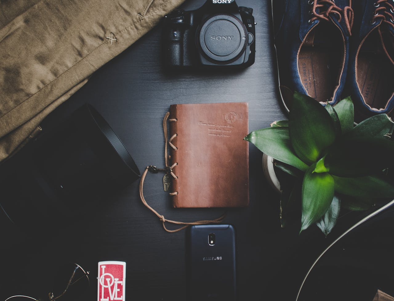 Flatlay featuring travel essentials including a camera, notebook, smartphone, and plant on a dark surface.